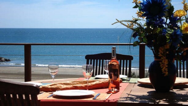 A dining table with a bottle, glasses, and a plate set on a wooden deck overlooking the ocean.