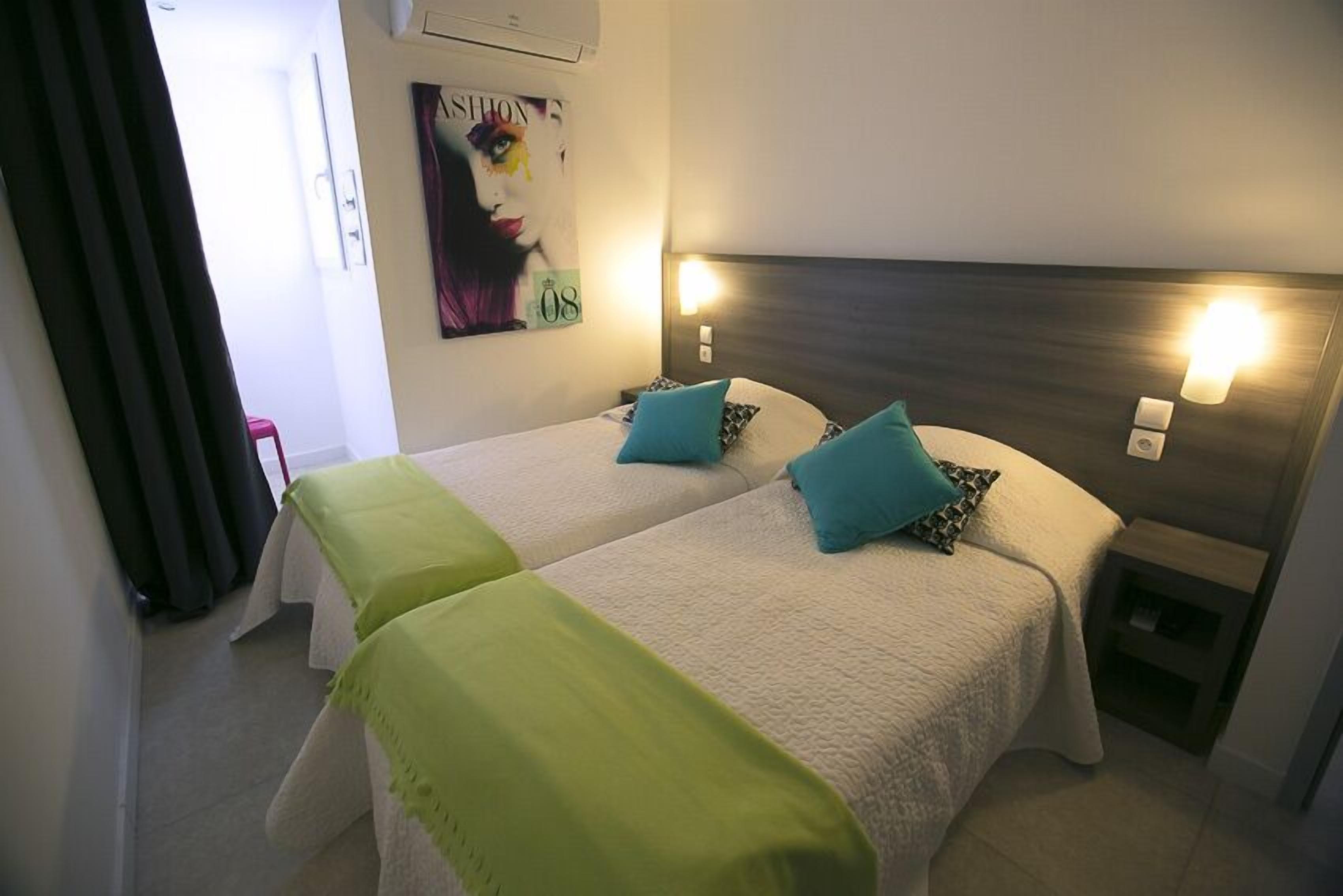 A hotel room with two beds, each with a headboard and bedside lamps. There is a small table and a chair in the corner.