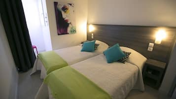 A hotel room with two beds, each with a headboard and bedside lamps. There is a small table and a chair in the corner.