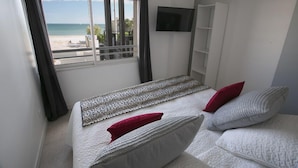 A bedroom with a bed, a TV, a window with a view of the beach, and a balcony.