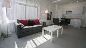A modern living room with a grey sofa, white coffee table, and a dining area with a small table and chairs. A kitchen with white cabinets and appliances is visible in the background.