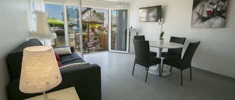 A modern living room with a sofa, a dining table with chairs, a TV mounted on the wall, and a sliding glass door leading to an outdoor area.