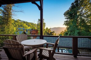 A wooden deck with a round table and chairs, overlooking a body of water and a forested hillside.