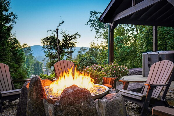 An outdoor seating area with a fire feature, surrounded by rocks and chairs, overlooking a natural landscape.