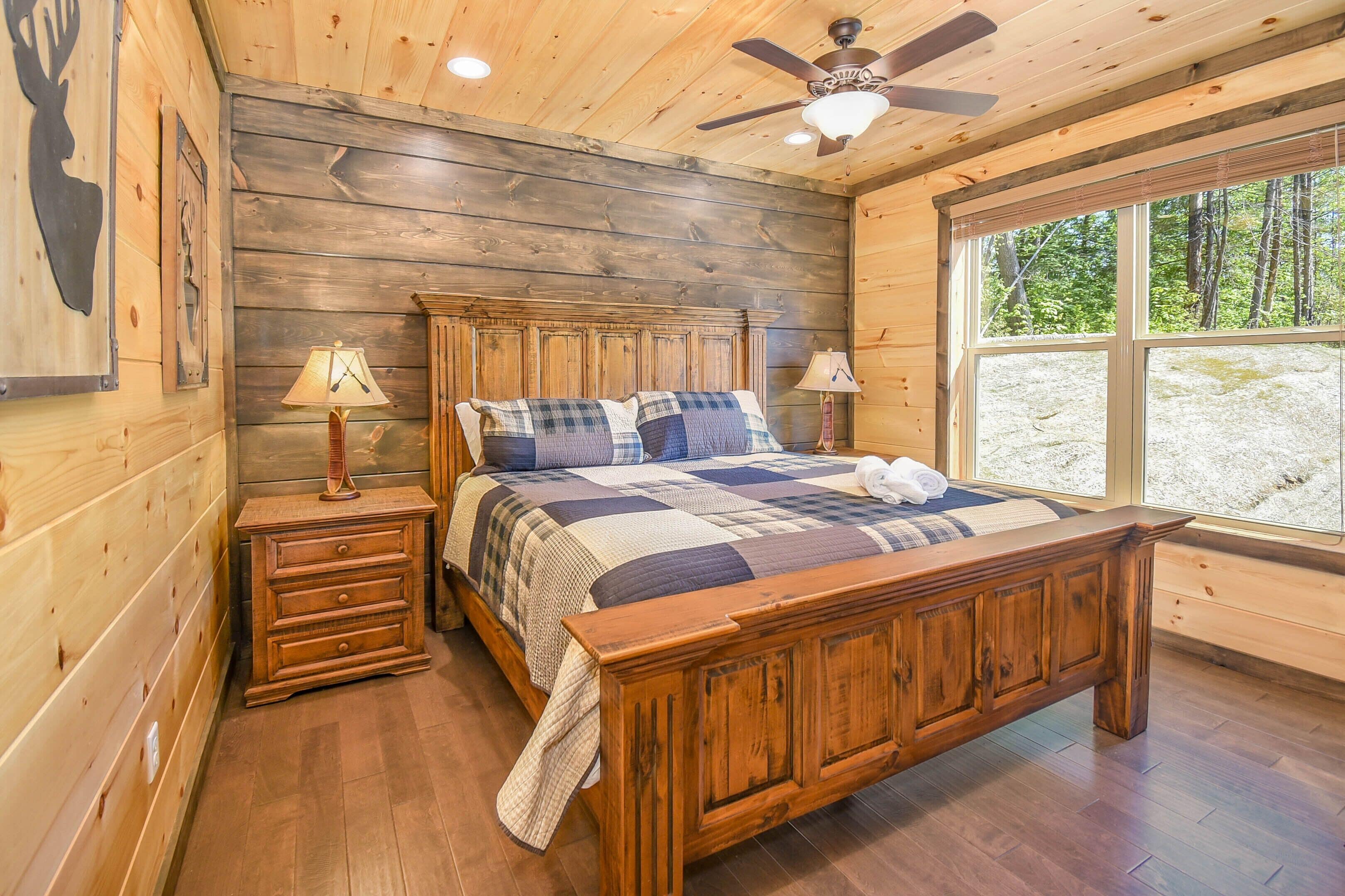 Un dormitorio de cabaña de madera con una cama grande, dos mesitas de noche, un ventilador de techo y una ventana con vistas al bosque.