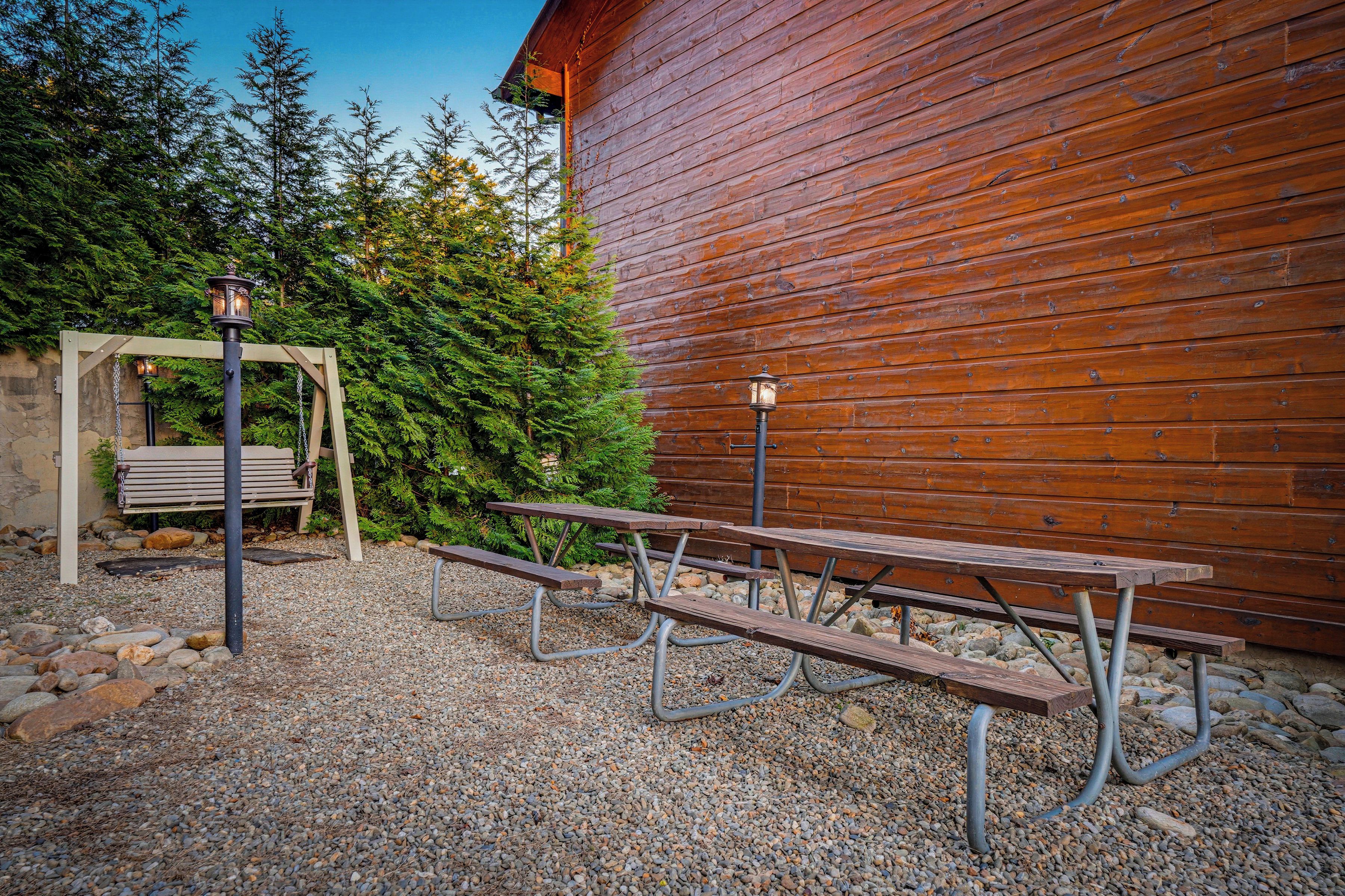 Un espace extérieur aménagé avec des tables de pique-nique et des bancs, un bâtiment en bois et, en arrière-plan, une rangée d’arbres.
