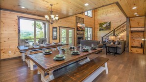 A spacious wooden interior with a large dining table set for a meal, a fireplace, and a staircase leading to an upper level.