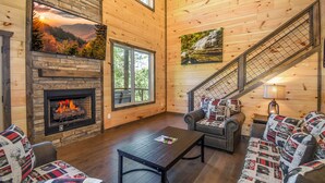 A cozy cabin interior with a fireplace, wooden walls, a staircase, and comfortable seating.