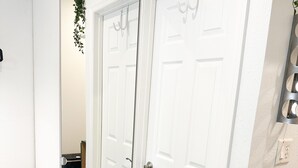 Un pasillo con puertas dobles blancas, un taburete pequeño, un espejo que refleja las puertas y un interruptor de luz en la pared.
