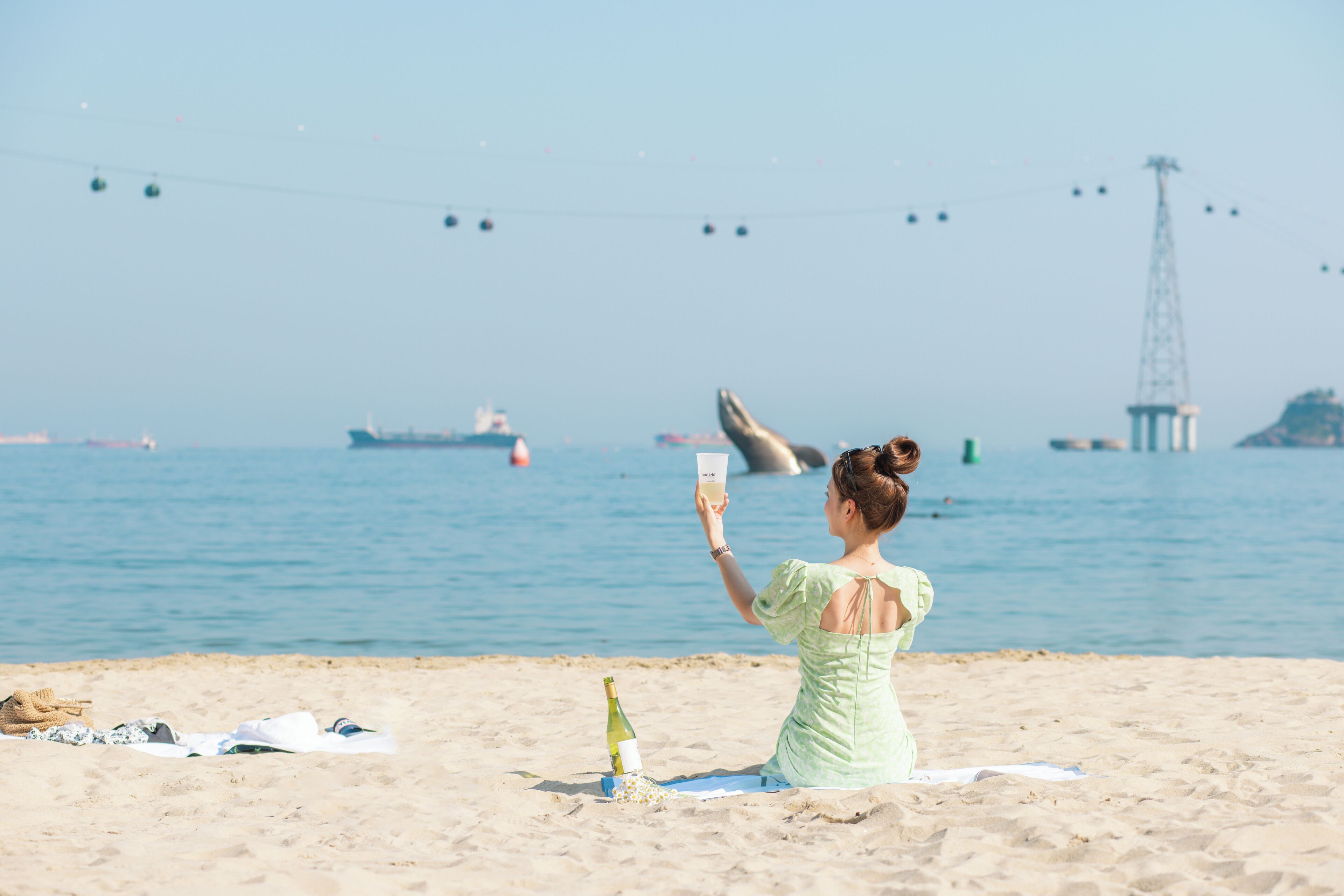 A woman sitting on a beach towel, holding a drink, with a bottle and clothes on the sand.