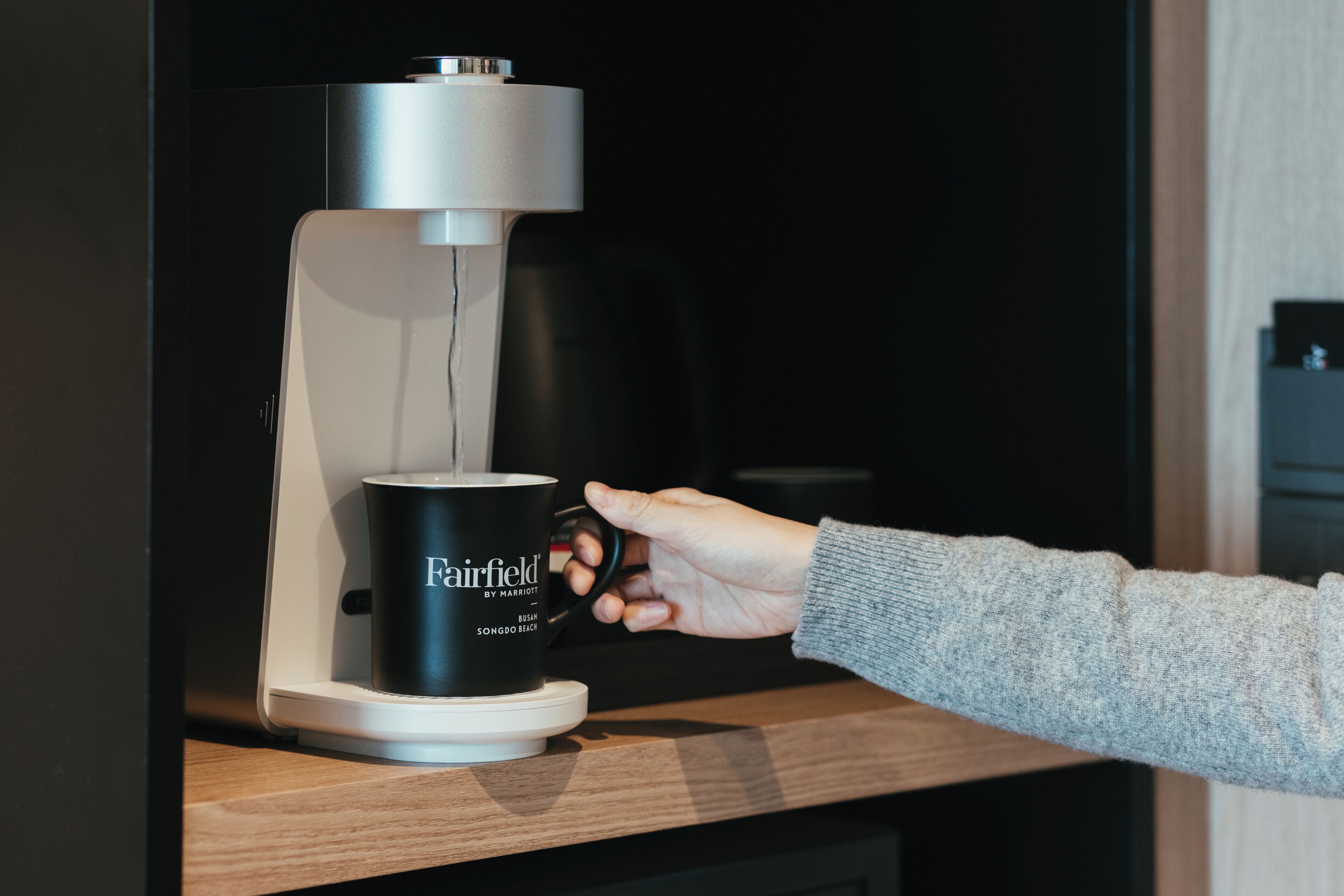 A hand holding a black mug labeled "Fairfield by Marriott" next to a coffee machine.