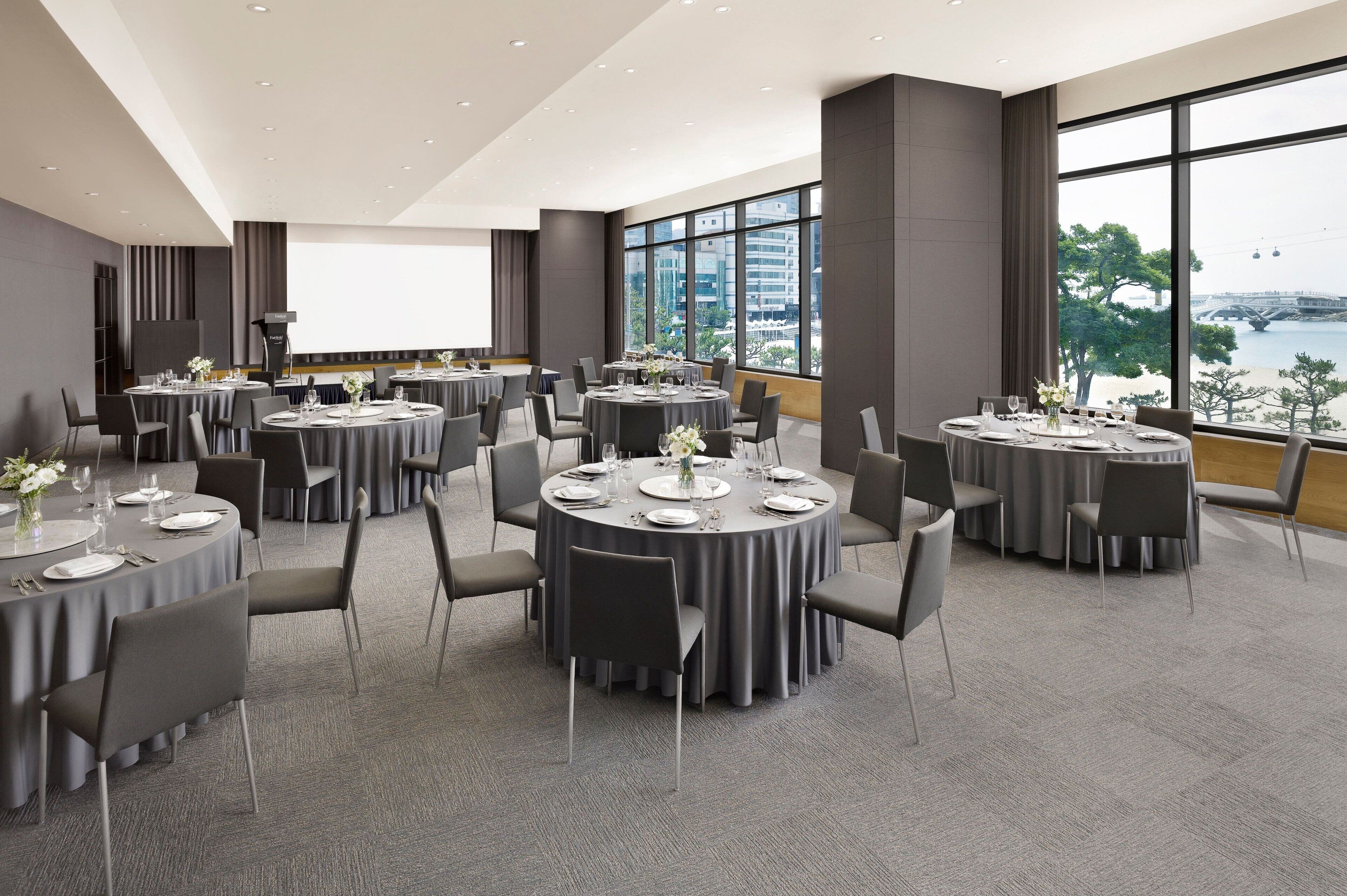 A modern conference room with round tables, grey chairs, and a large window offering a view of a cityscape.