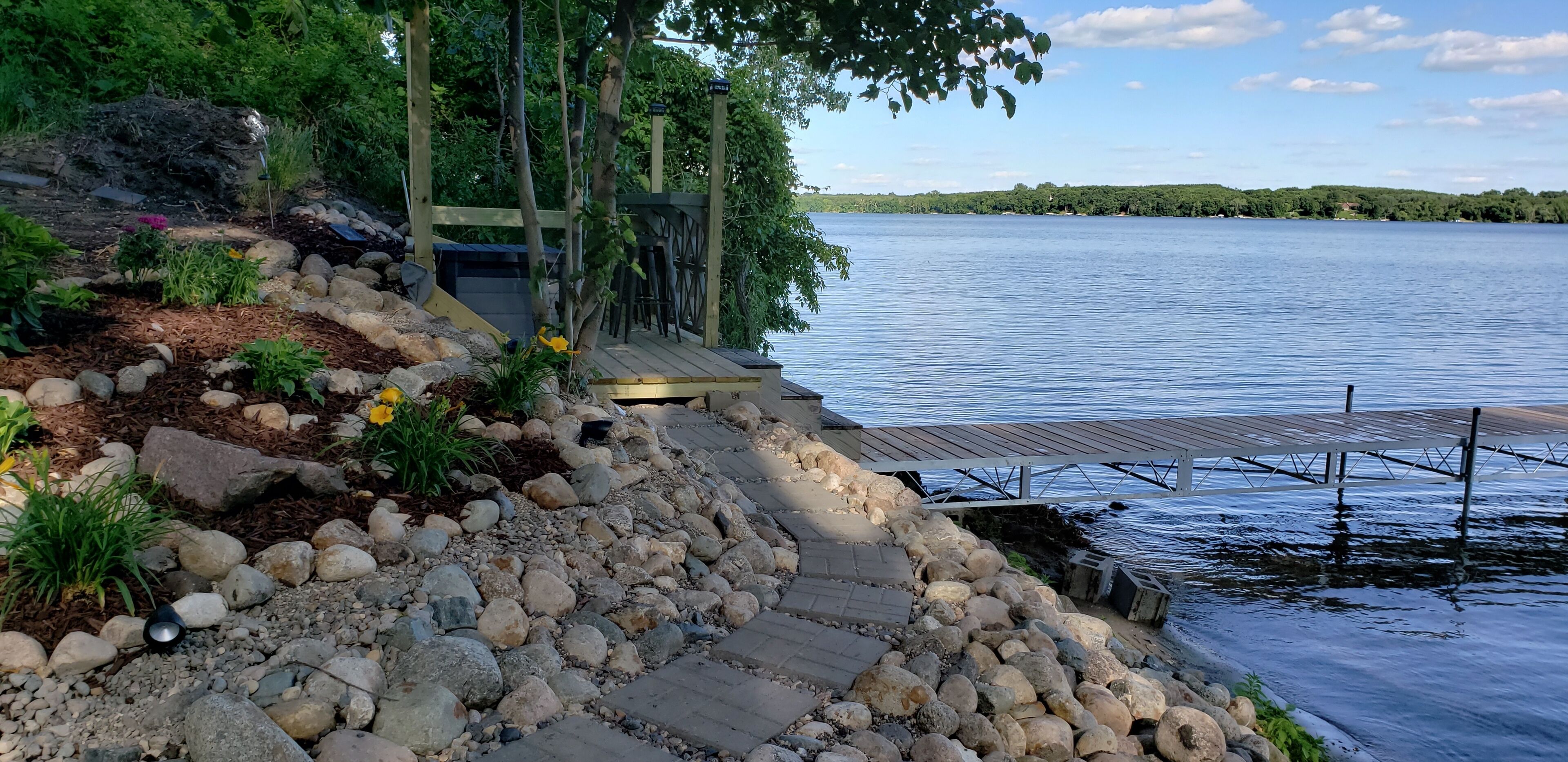 A wooden dock extending into a calm lake, surrounded by a rocky shoreline with a wooden deck and a small structure.