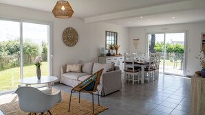 A spacious living room with a large dining table, a sofa, and a coffee table. There is a large window providing a view of the garden.