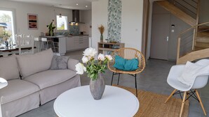 A modern living room with a sofa, a round table, and a wicker chair. A staircase leads to an upper level.
