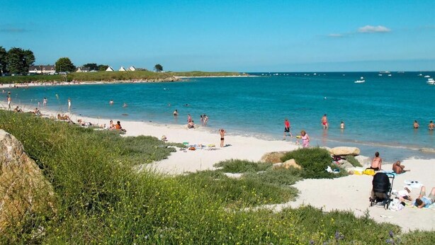 A beach with people sunbathing and swimming, a clear blue sky, and boats in the water.