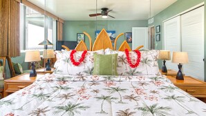 A hotel room with a bed featuring a floral and palm tree bedspread, two nightstands with lamps, a ceiling fan, and a view of the outside through a window with curtains.