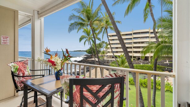 A balcony with a table and chairs, overlooking a beach with palm trees and a hotel building.
