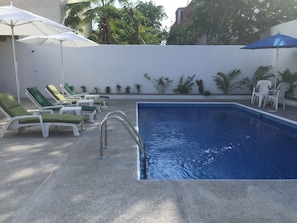 A swimming pool with sun loungers and umbrellas, surrounded by plants and a white wall.
