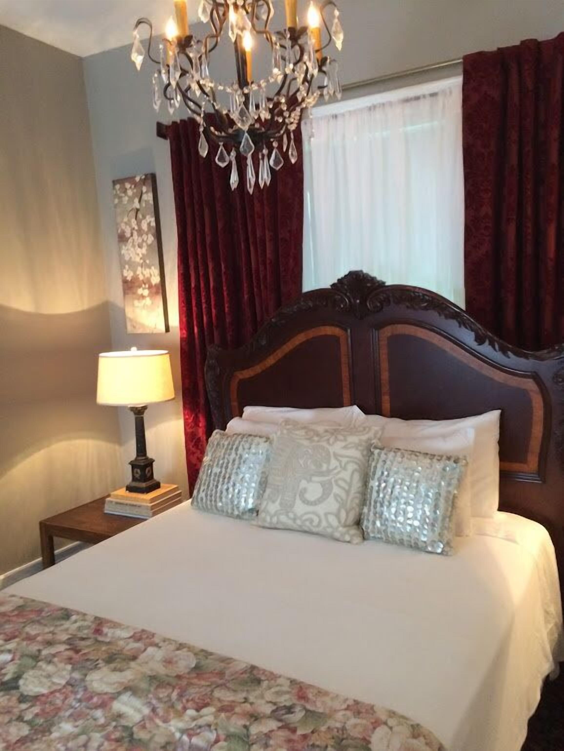 A bedroom with a large bed, a chandelier, a nightstand with a lamp, and red curtains.