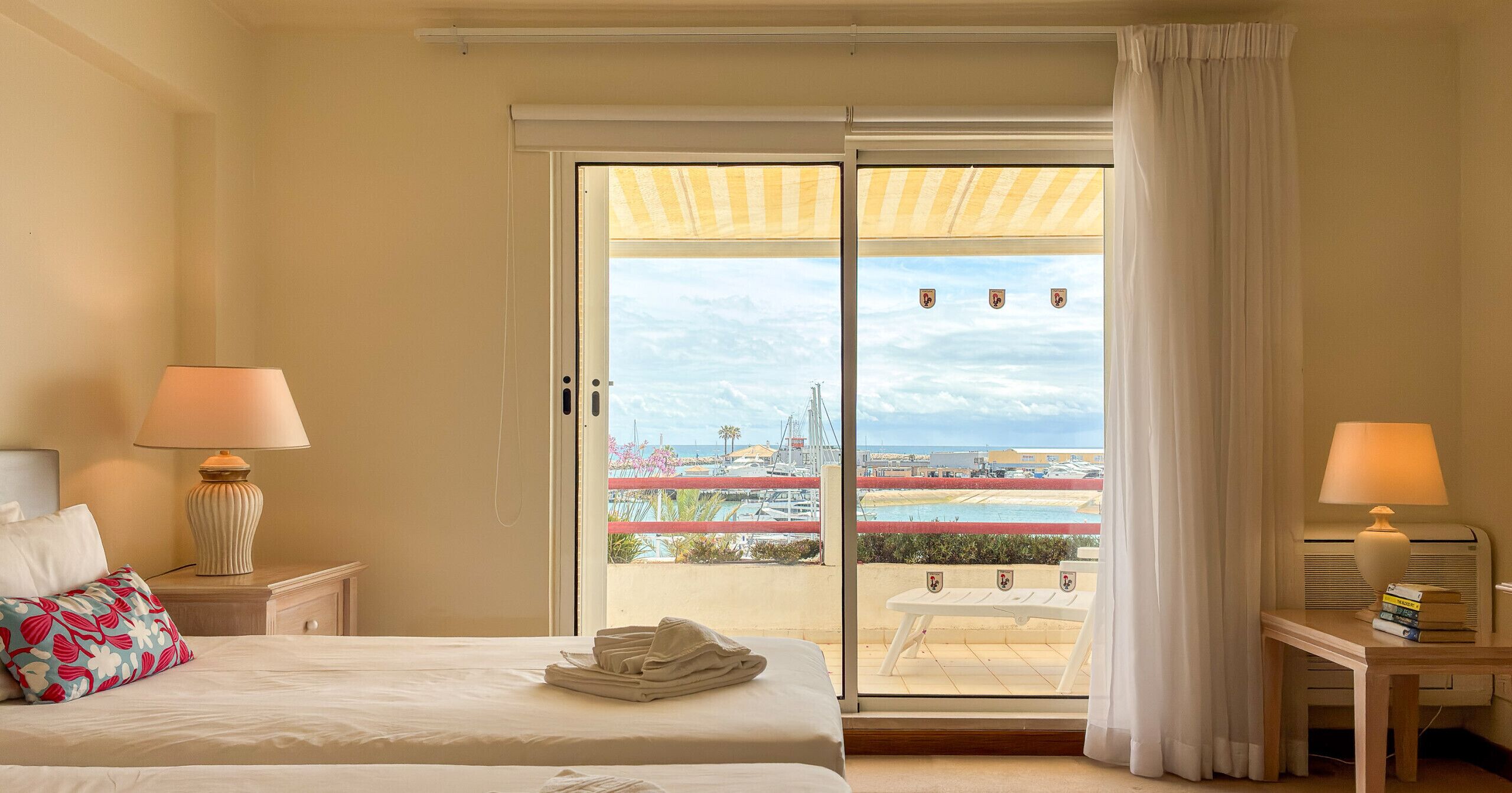 Une chambre d’hôtel avec un lit, deux lampes de chevet, une table avec des livres et une vue sur un port de plaisance, visible à travers une porte-fenêtre coulissante.
