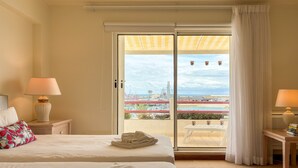 Habitación de hotel con cama, dos lámparas de noche, una mesa con libros y vistas a un puerto deportivo a través de una puerta corrediza de vidrio.