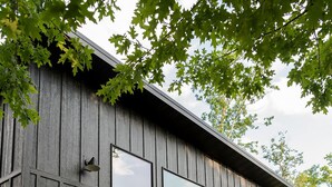 Una cabaña moderna de madera con ventanas amplias, una terraza con mobiliario exterior y un entorno boscoso.