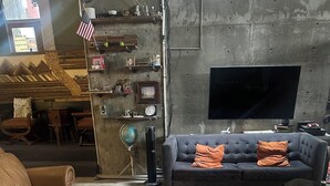 Una sala de estar con un sofá de cuero, un sofá gris con cojines naranja, una mesa de centro de madera y un televisor montado en una pared de concreto.