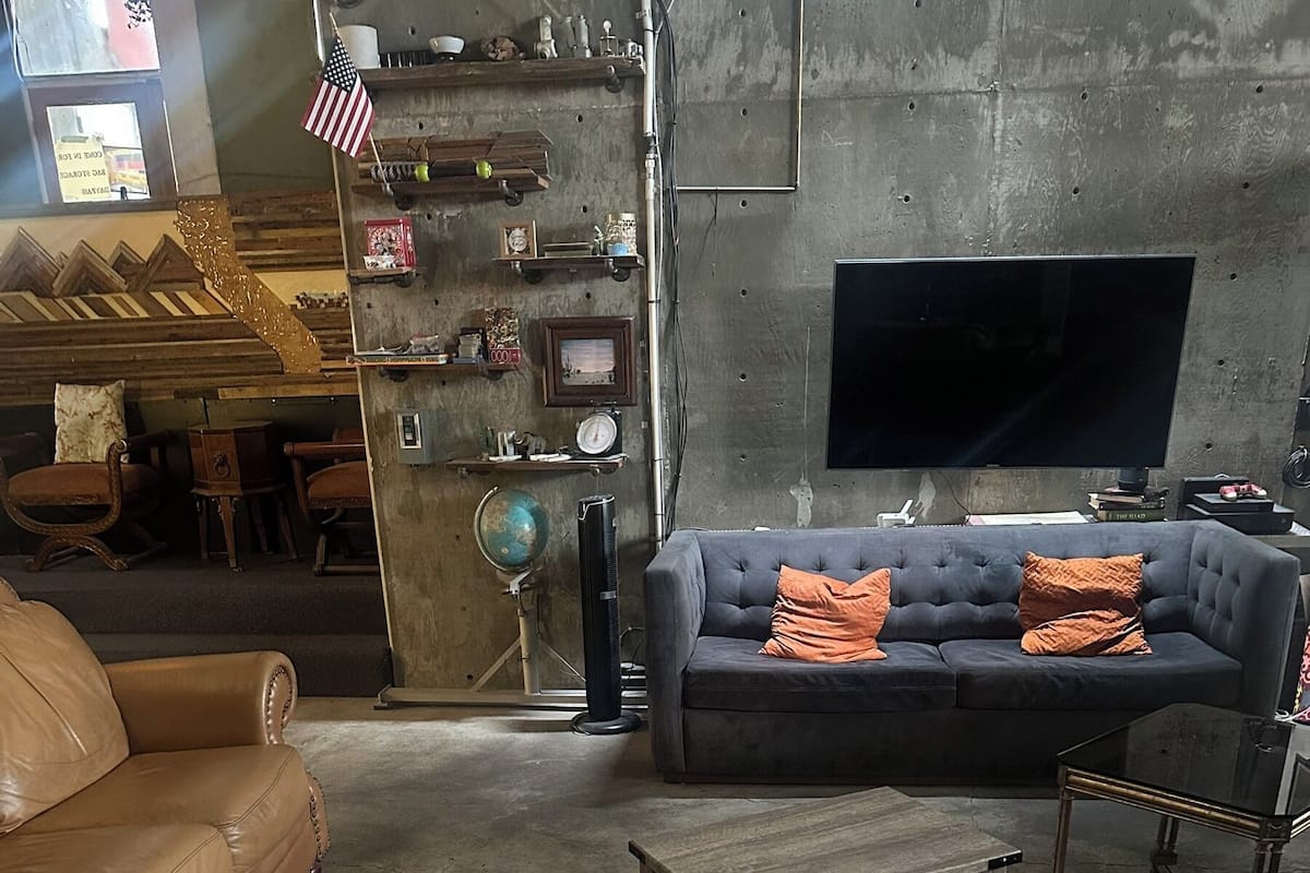 Un salon avec un canapé en cuir, un canapé gris agrémenté de coussins orange, une table basse en bois et un téléviseur fixé à un mur en béton.