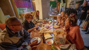 Un grupo de personas comiendo en una mesa de madera, con diferentes platillos y una bolsa de bocadillos orgánicos.
