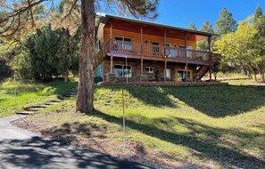 A two-story log cabin with a balcony, surrounded by trees and a grassy area.