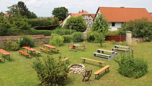 Un espace extérieur aménagé en terrasse, avec plusieurs tables et bancs en bois, entouré de verdure et agrémenté d’un foyer en pierre.