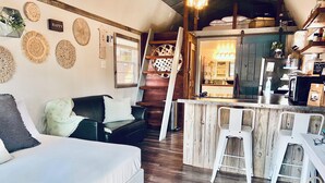 A cozy living area with a sofa, a small kitchen area with a bar, and a staircase leading to an upper level. - Private & Cozy cabin with Hot Tub-Coffee Bar-Cowboy Pool (Hot Springs)