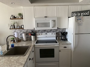 Una cocina moderna con armarios blancos, un microondas integrado, una estufa y un refrigerador. Hay una estantería con objetos decorativos y un cartel que dice 'life is good'.