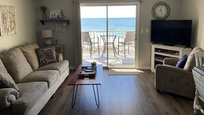 A living room with a sofa, a coffee table, a TV, and a view of the ocean through a sliding glass door.