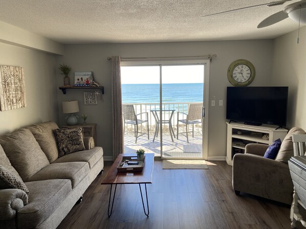 A living room with a sofa, a coffee table, a TV, and a view of the ocean through a sliding glass door.