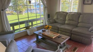 Un salon avec un canapé gris, une table basse sur laquelle se trouve un plateau, et une vue sur un paysage verdoyant par de grandes fenêtres.