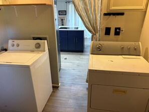 A laundry room with two front-loading washers and dryers, a wooden floor, and a view into the kitchen area.