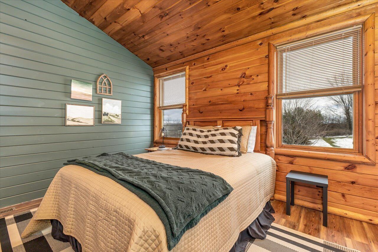 A cozy bedroom with a wooden ceiling, a bed with a quilted blanket, two bedside tables, and a window with blinds.