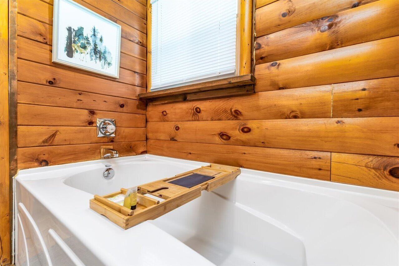 A wooden cabin bathroom with a bathtub, a wooden tray with toiletries, a framed picture on the wall, and a window with blinds.