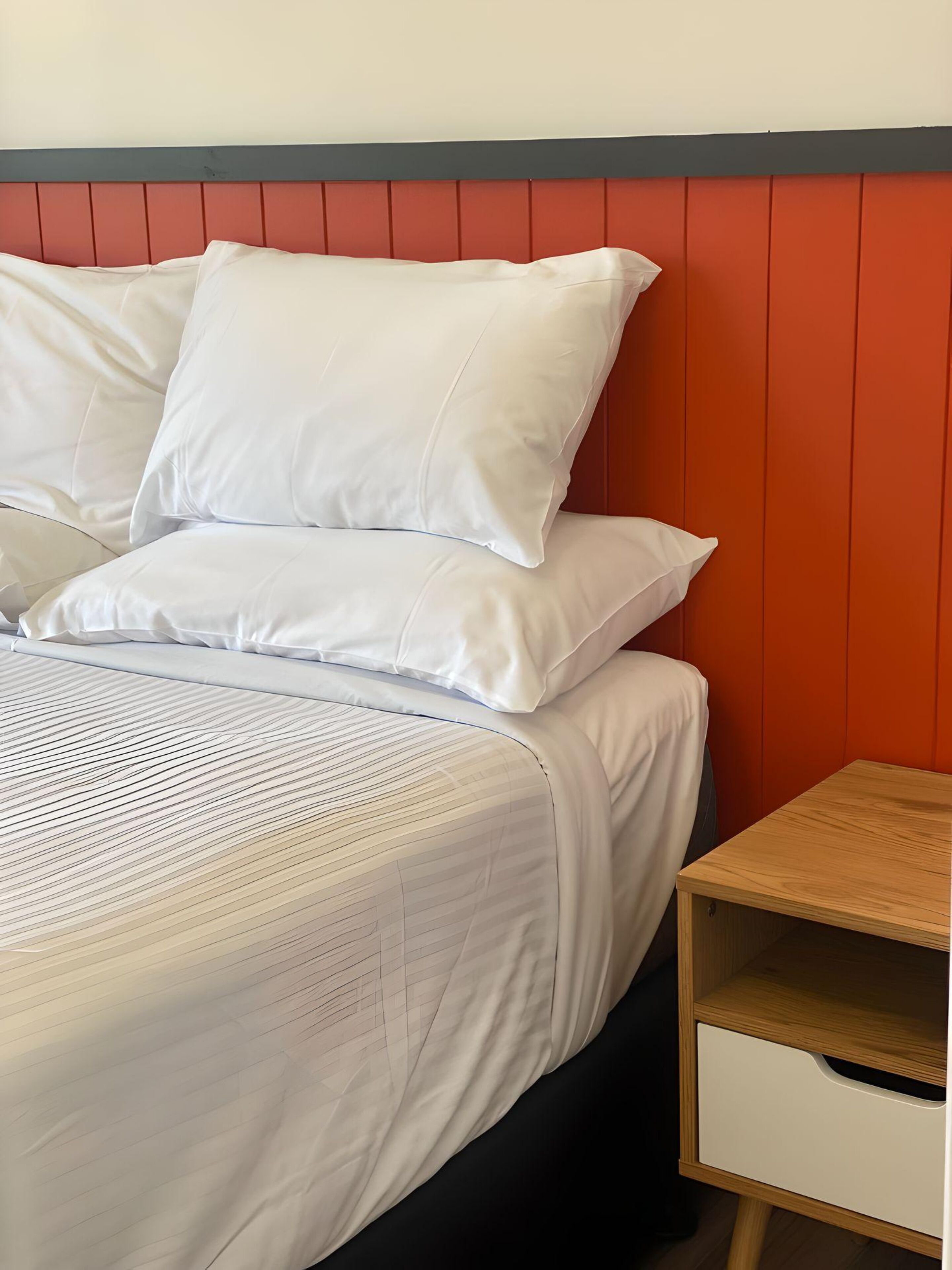 Een bed met witte beddengoed en een rood hoofdeinde, een houten nachtkastje met een lade, en een effen muur.
