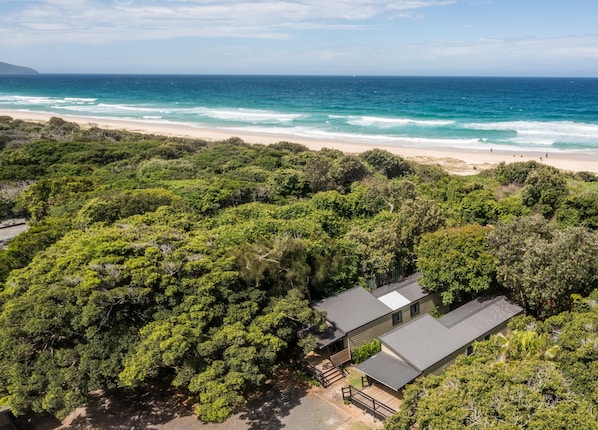 An aerial view of a coastal property with dense greenery, a beach, and a small building with a sloped roof. - Tiona Palms Holiday Park (Tiona)