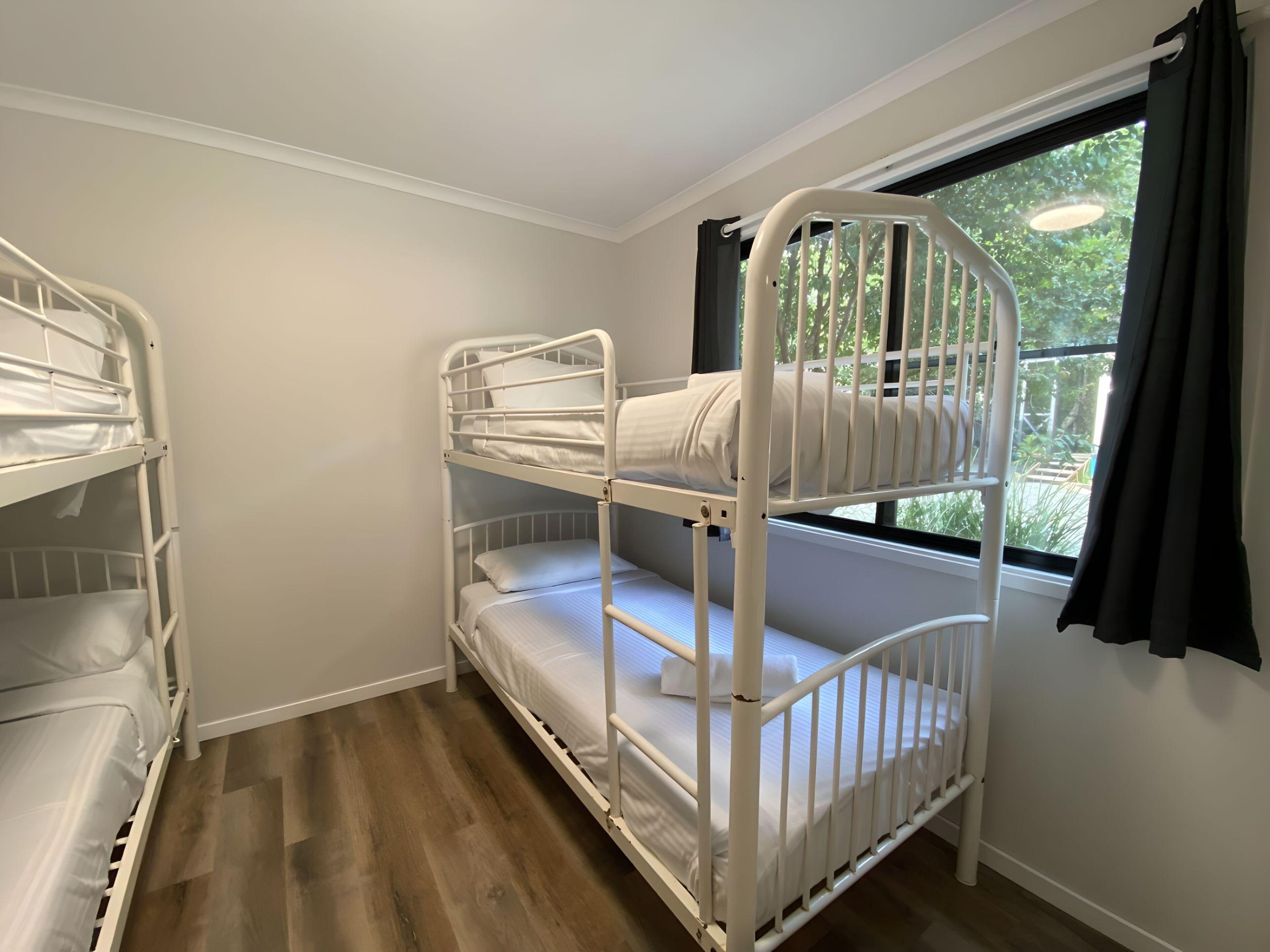 A bunk bed room with a window and curtains, wooden flooring, and white walls.