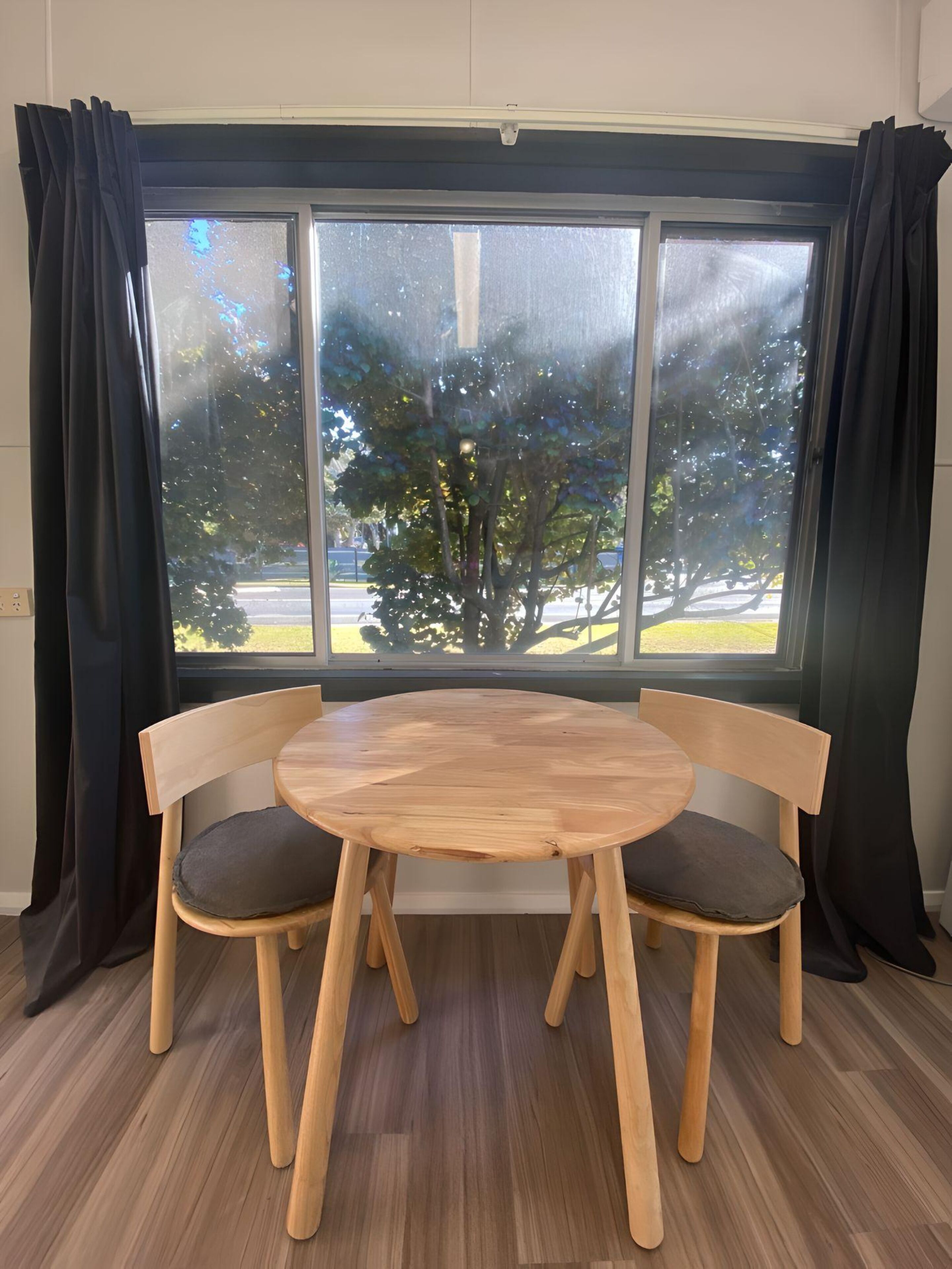 A room with a round wooden table and two chairs, a large window with black curtains, and a view of trees and a road outside.