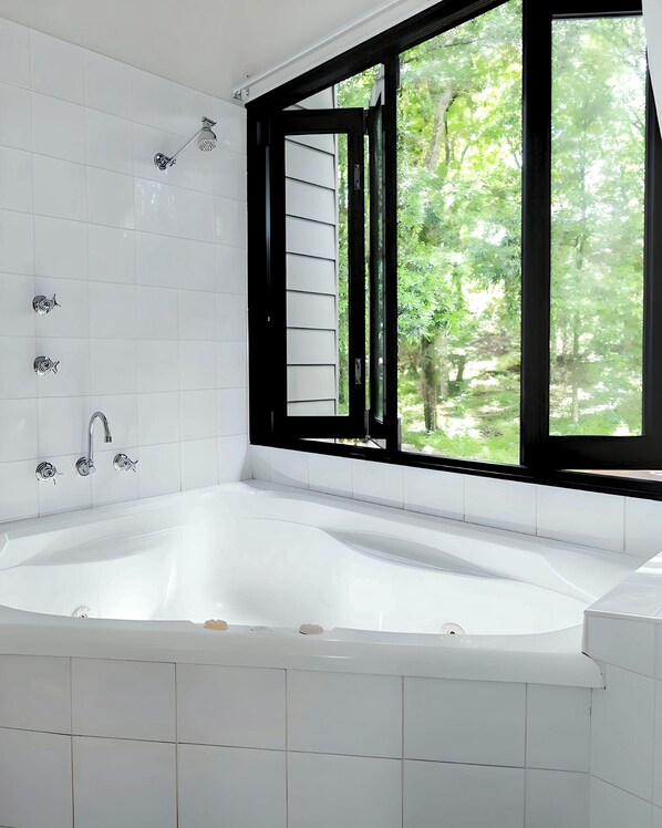 A modern bathroom with a large bathtub, white tiles, a window with black frames, and a view of greenery outside. - Tiona Palms Holiday Park (Tiona)