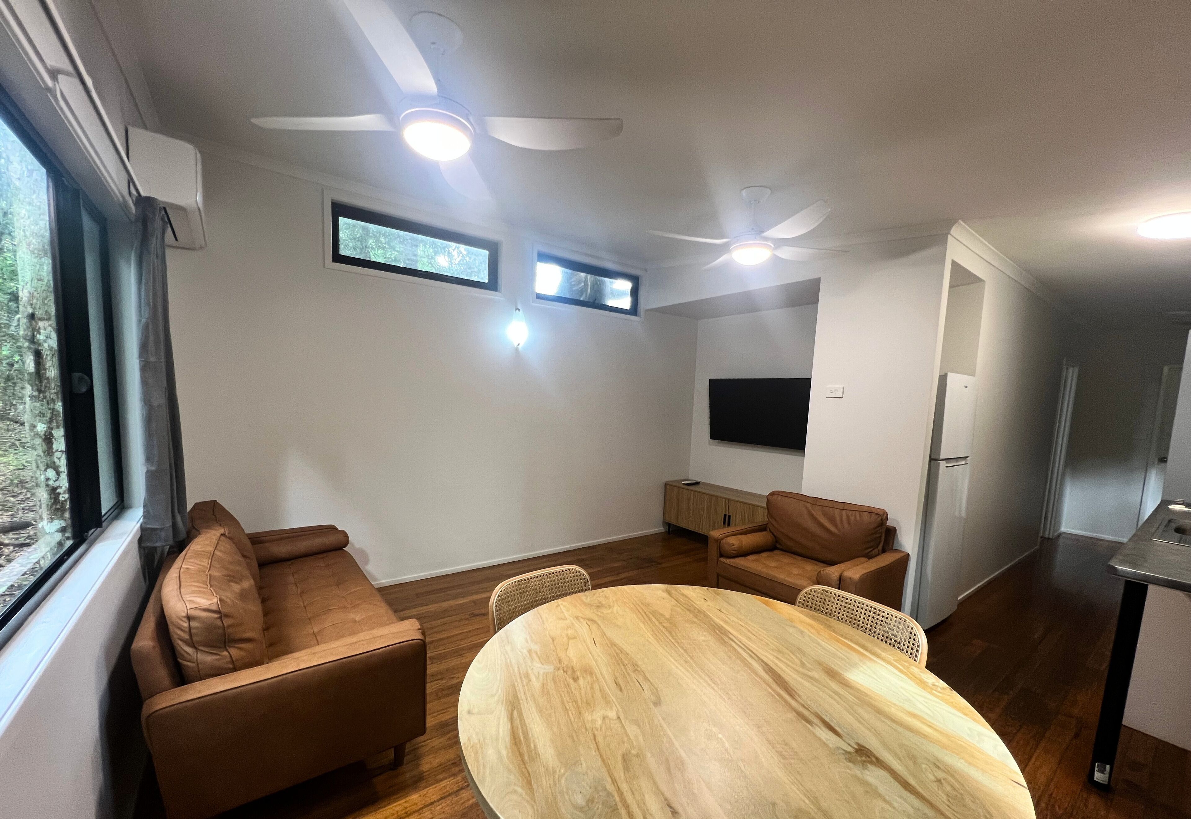 Un salon moderne avec une table ronde en bois, deux canapés marron, des ventilateurs de plafond et un téléviseur à écran plat fixé au mur.