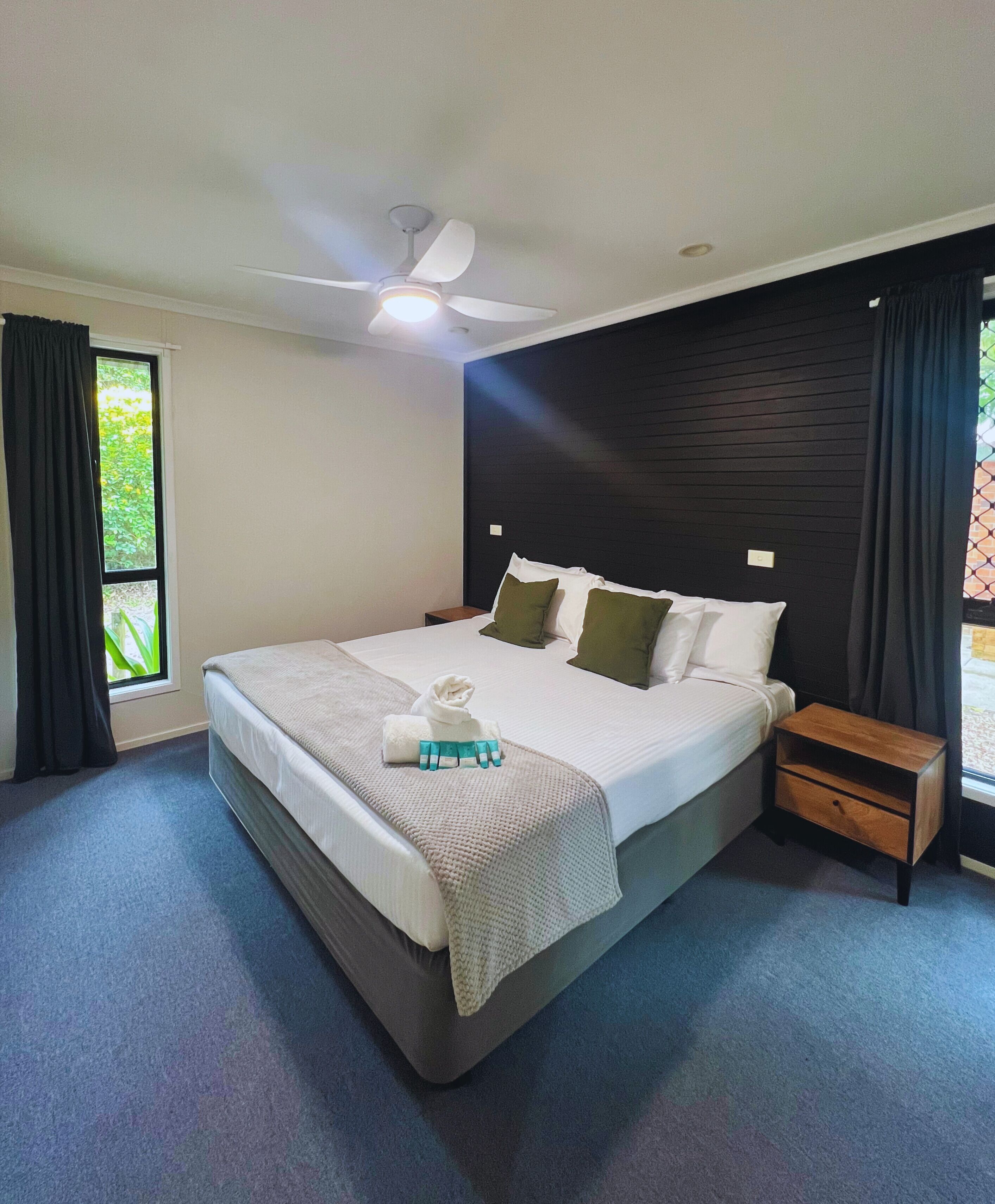 A bedroom with a large bed, a nightstand, a ceiling fan, and a window with curtains.