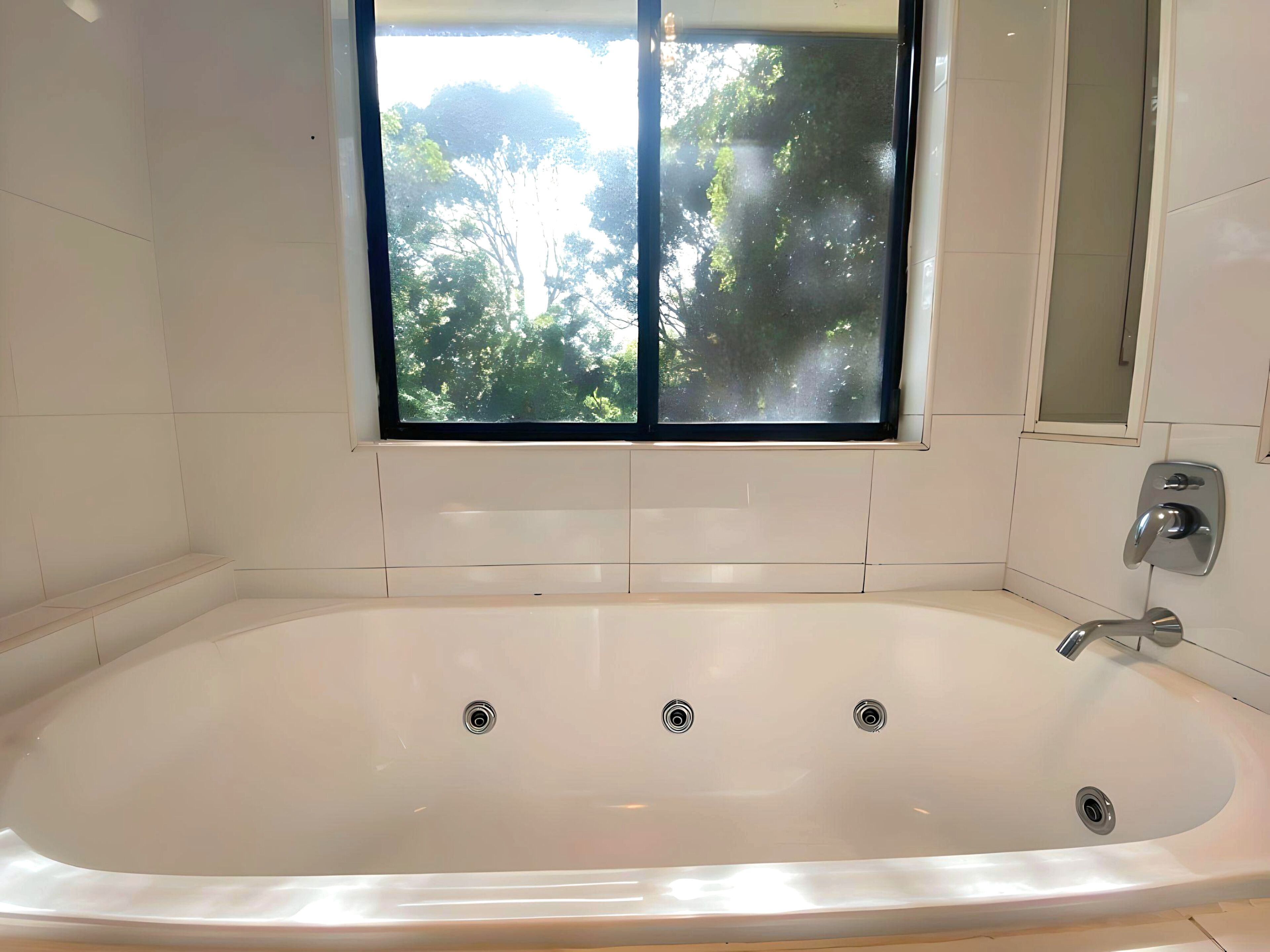 A modern bathroom with a large bathtub, a window with a view of trees, and a mirror on the wall.