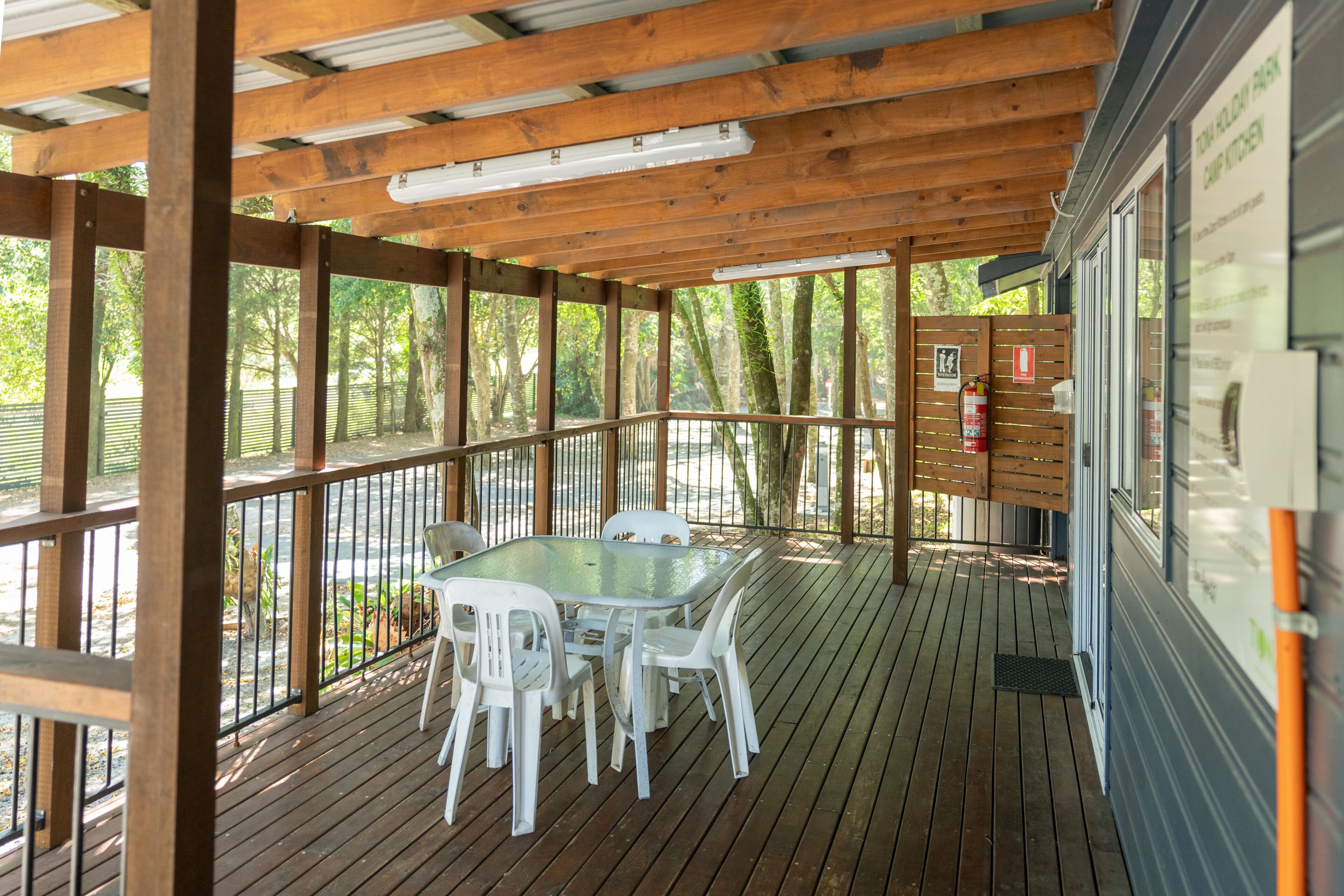 A covered outdoor area with a wooden deck, a glass table with chairs, and a view of trees.