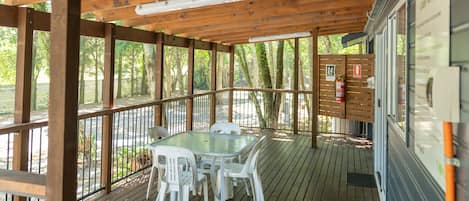 A covered outdoor area with a wooden deck, a glass table with chairs, and a view of trees.